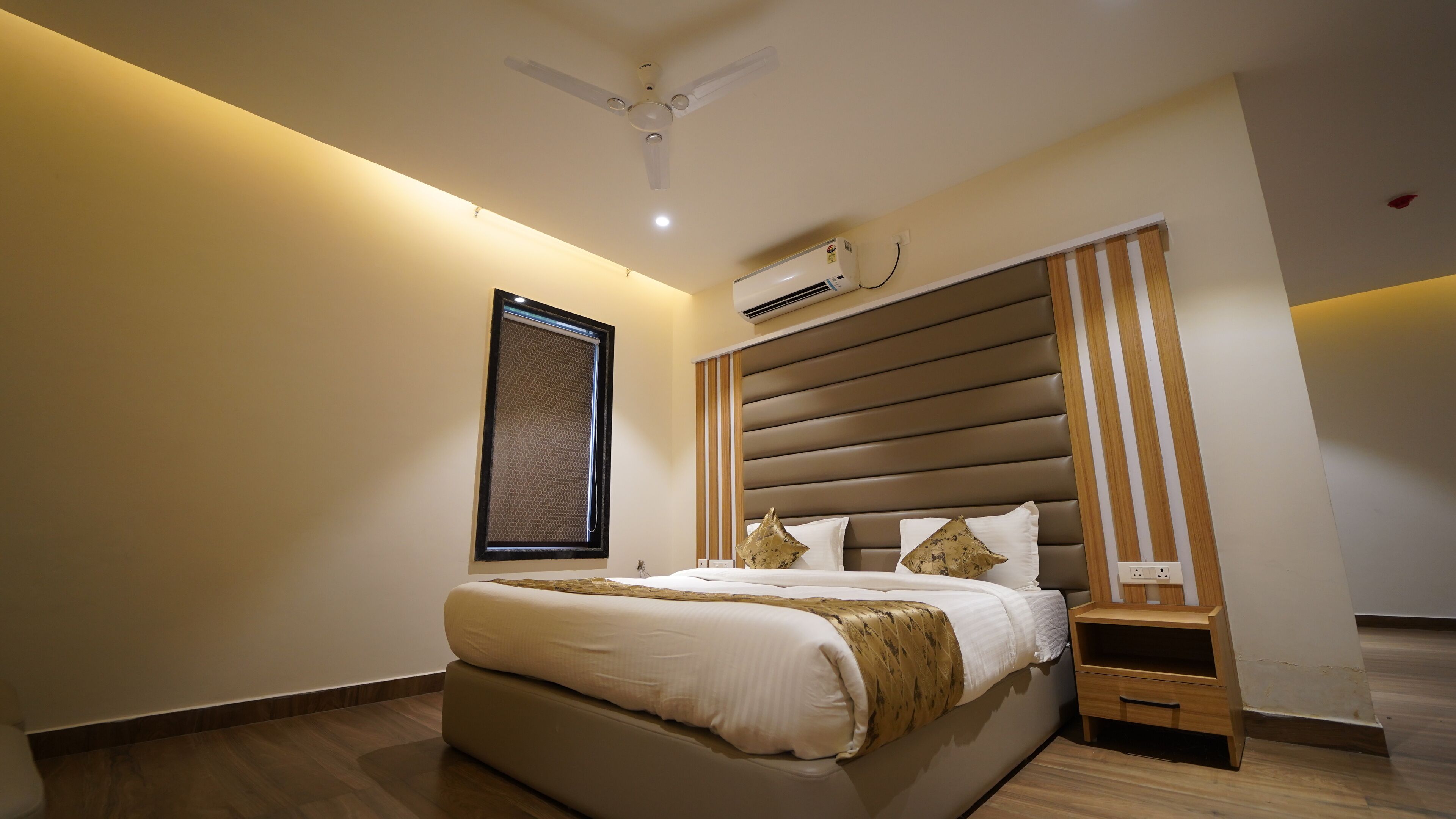 Un dormitorio moderno con una cama grande, un ventilador de techo, un equipo de aire acondicionado, una mesita de noche de madera y un cuadro enmarcado en la pared.