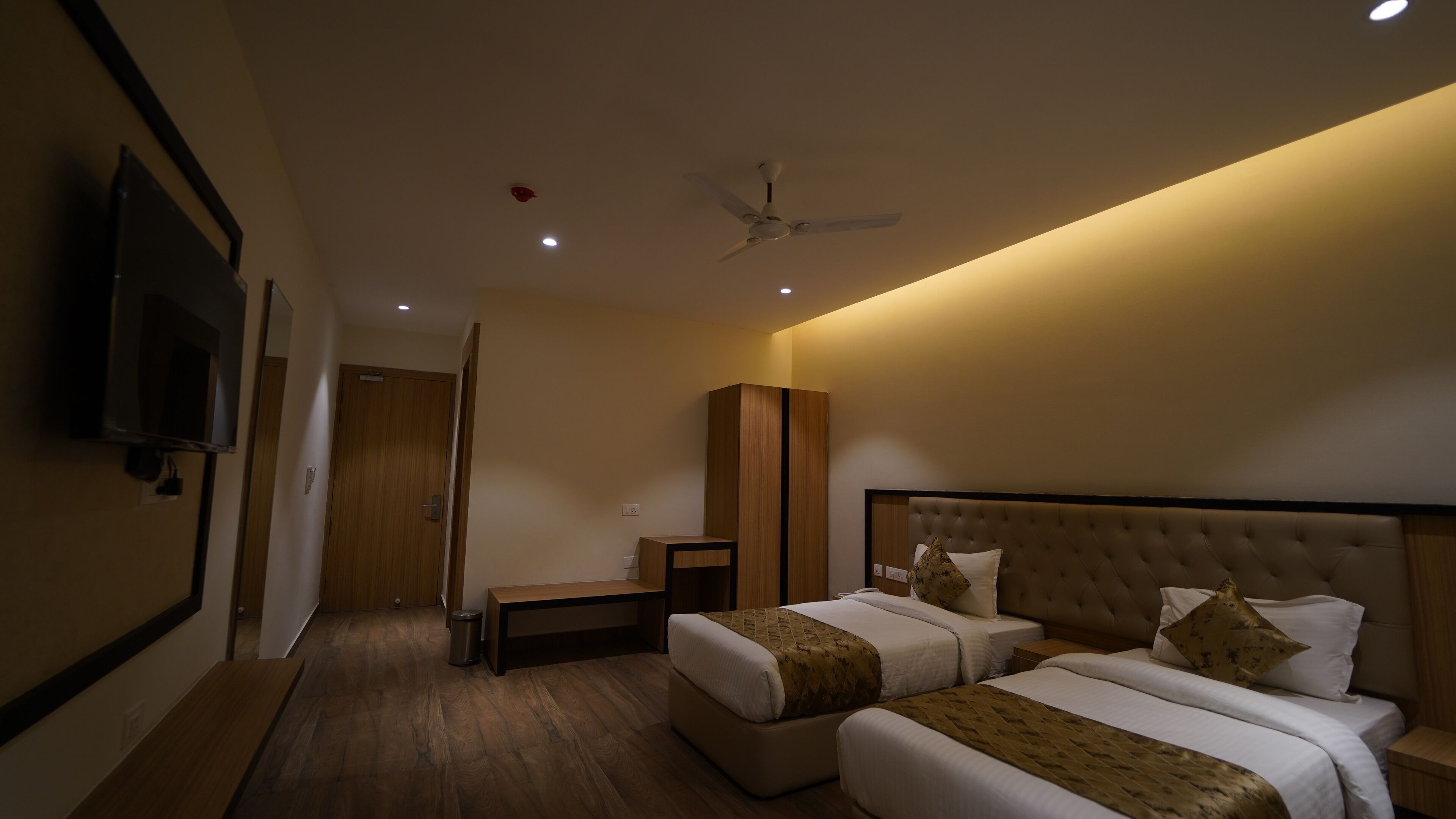 A hotel room with two beds, a TV mounted on the wall, a ceiling fan, and a wooden cabinet.