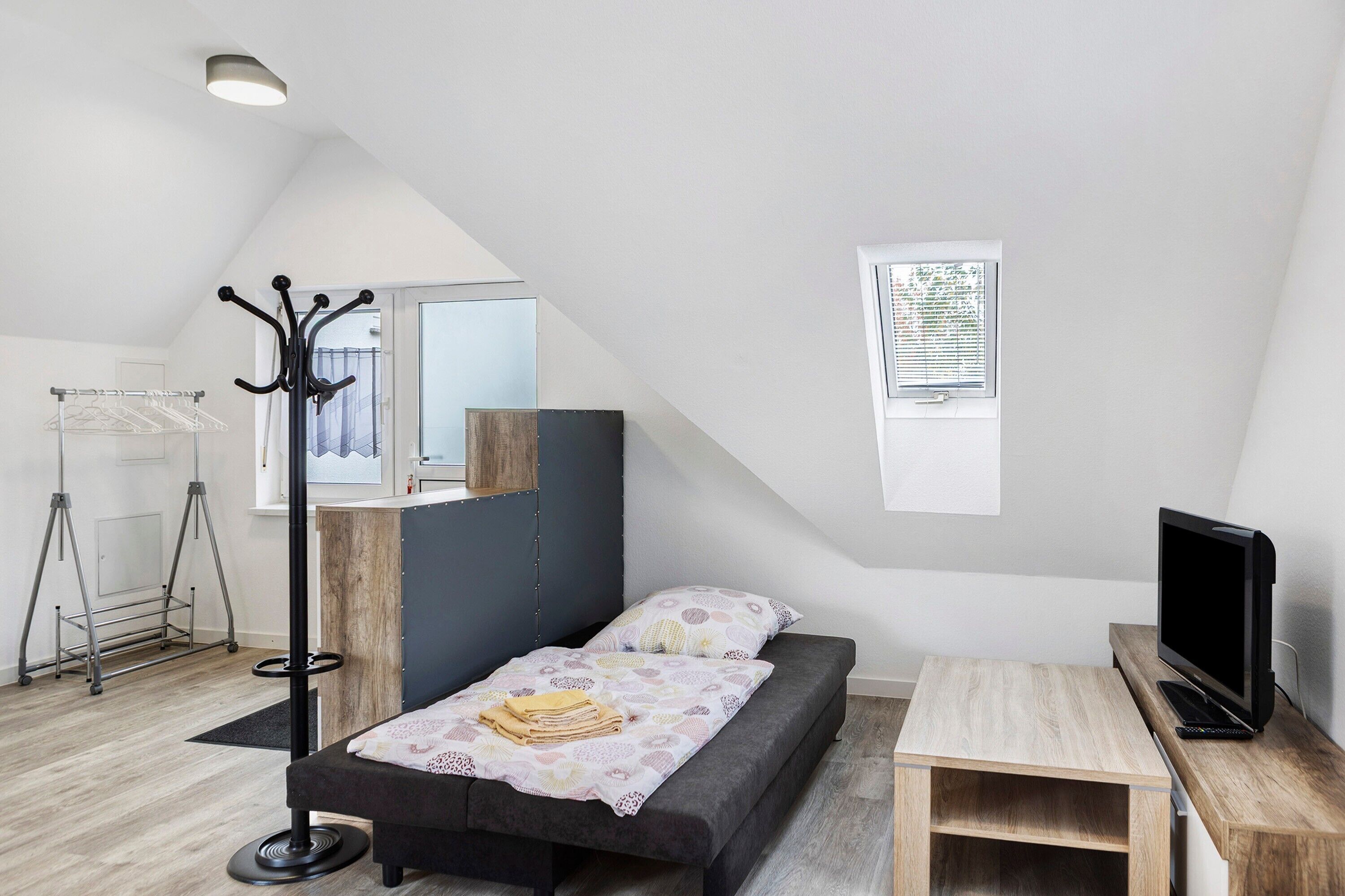 A bedroom with a bed, a TV, a desk, a coat rack, and a window.