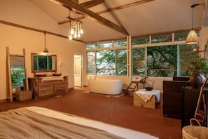 A spacious room with a large bed, a bathtub, and a wooden dresser. There are large windows providing natural light and views of trees.