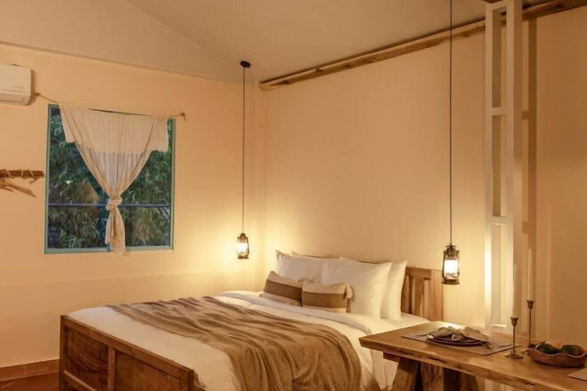 A bedroom with a bed, bedside table, hanging lanterns, and a window with a view.