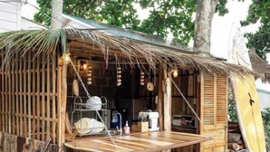 A rustic outdoor kitchen with a wooden table, hanging lights, and a surfboard leaning against the wall. - Canaan Beach Home (Phu Quoc Island)