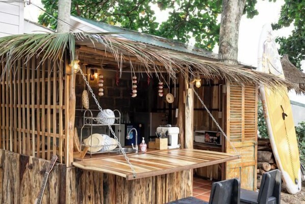A rustic outdoor kitchen with a wooden table, hanging lights, and a surfboard leaning against the wall. - Canaan Beach Home (Phu Quoc Island)