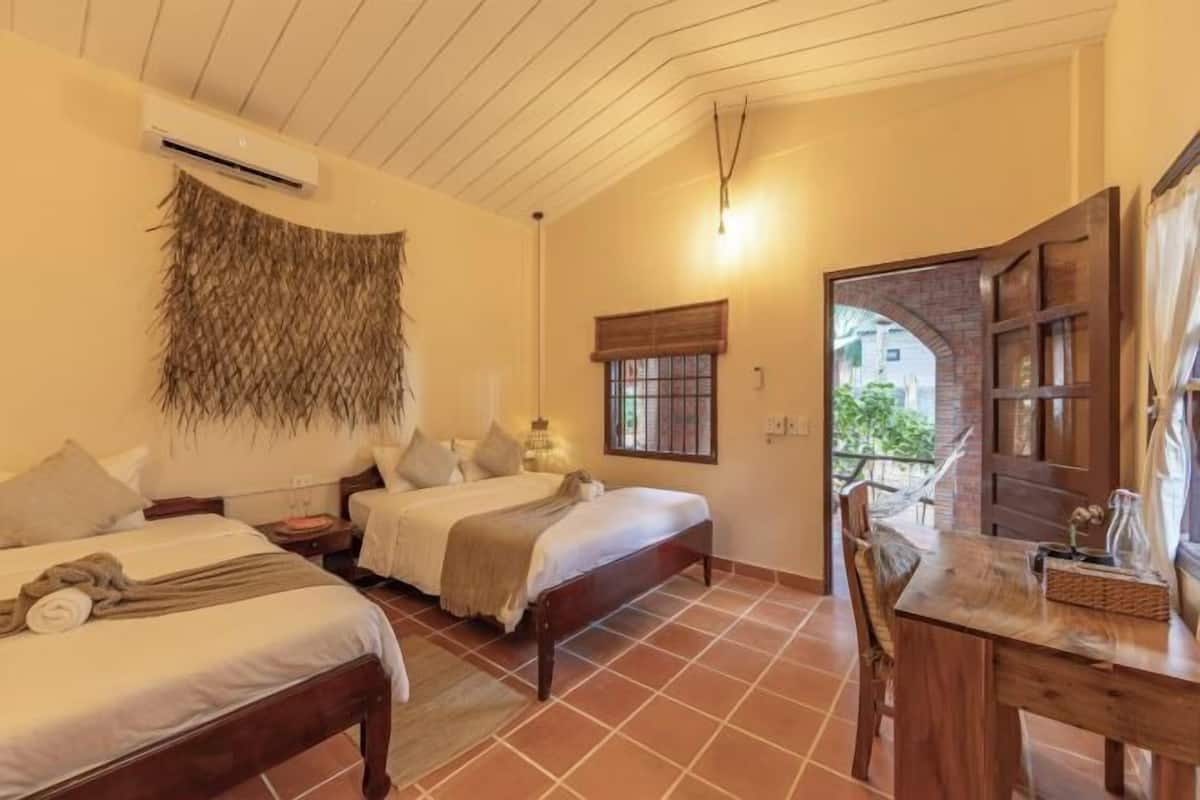 A bedroom with two beds, a wooden table, and a window with a view of greenery.
