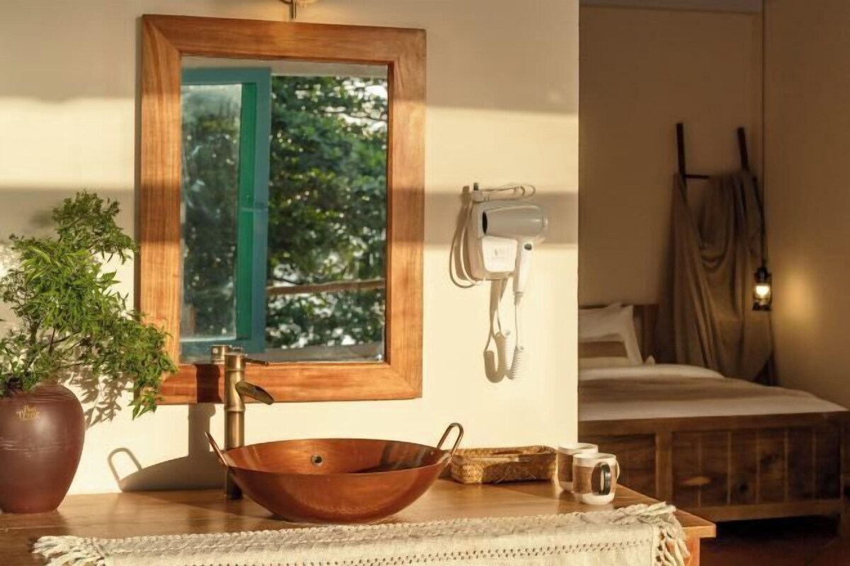 A room with a wooden-framed mirror, a copper basin, a potted plant, a bedside table with a mug, and a bed with a headboard.
