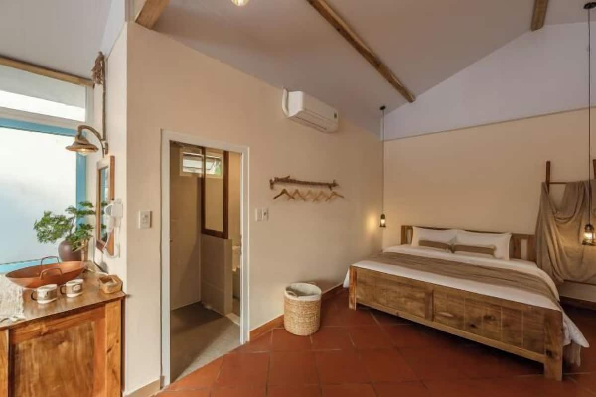 A bedroom with a wooden bed, a small bedside table, a hanging light fixture, and a window with a view.