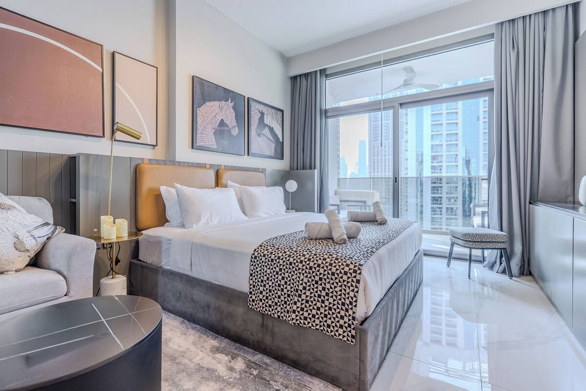 A modern hotel room with a large bed, a sofa, a small table, and a view of the city through large windows.