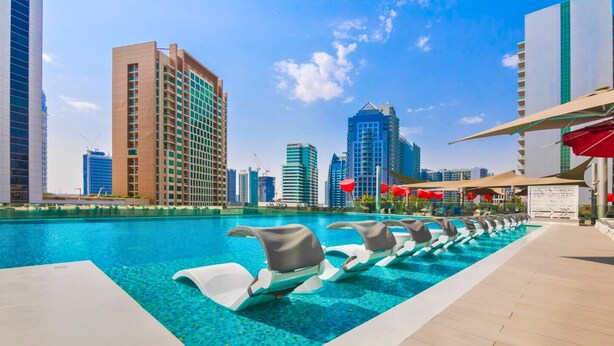 A swimming pool with lounge chairs, surrounded by high-rise buildings and a clear sky. - MAG 318 Residences - Business Bay- Dubai (Dubai)