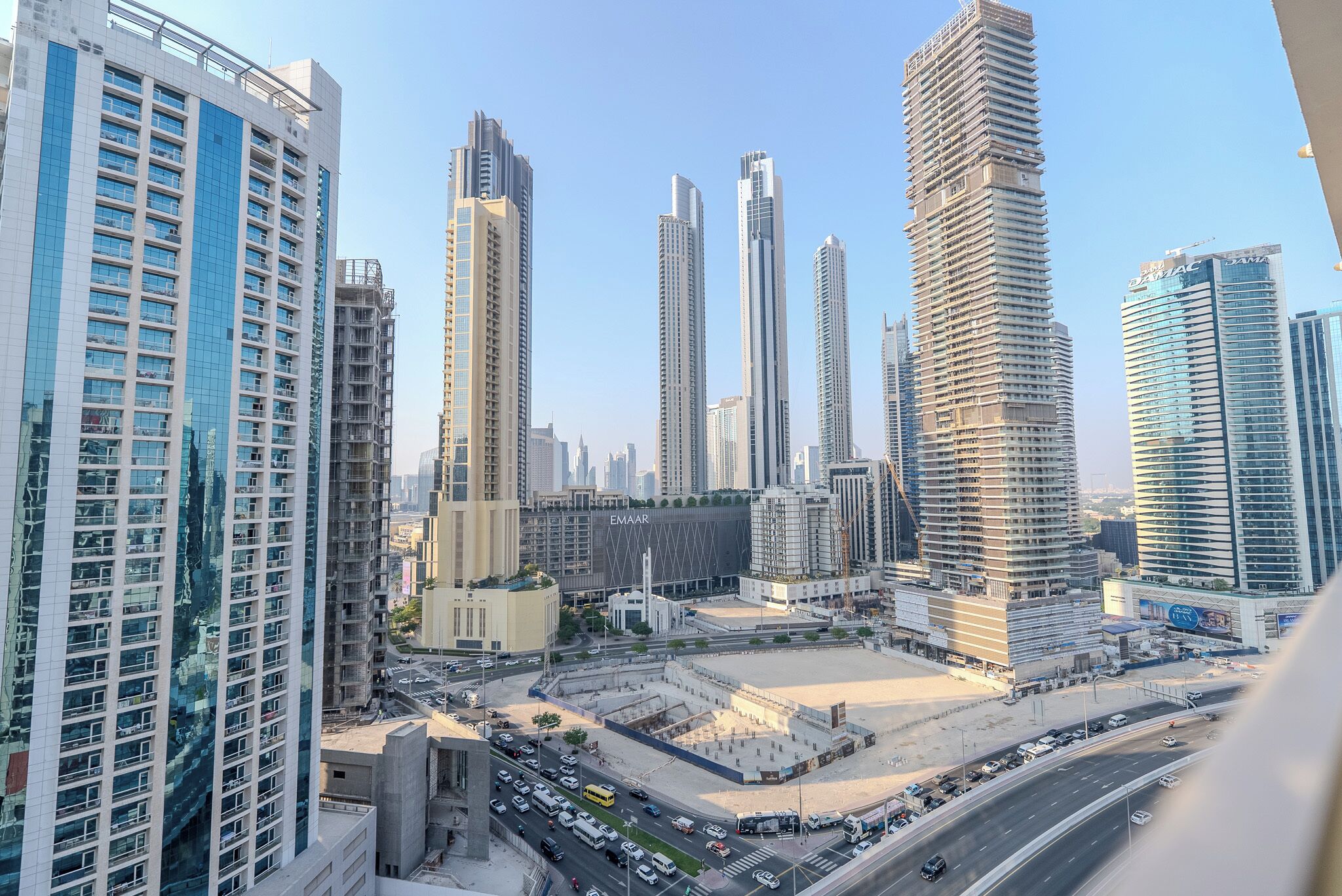 Uma área urbana com edifícios altos e modernos, uma ampla praça e uma rua movimentada com veículos.