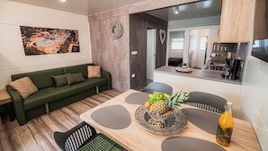 A modern living room with a green sofa, a dining table with a fruit centerpiece, and a kitchen area visible through an open door.