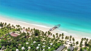 Luftaufnahme eines Strandes mit Palmen, einem Steg, der ins Wasser ragt, und einem Hotelbereich mit Gebäuden und Sonnenschirmen.