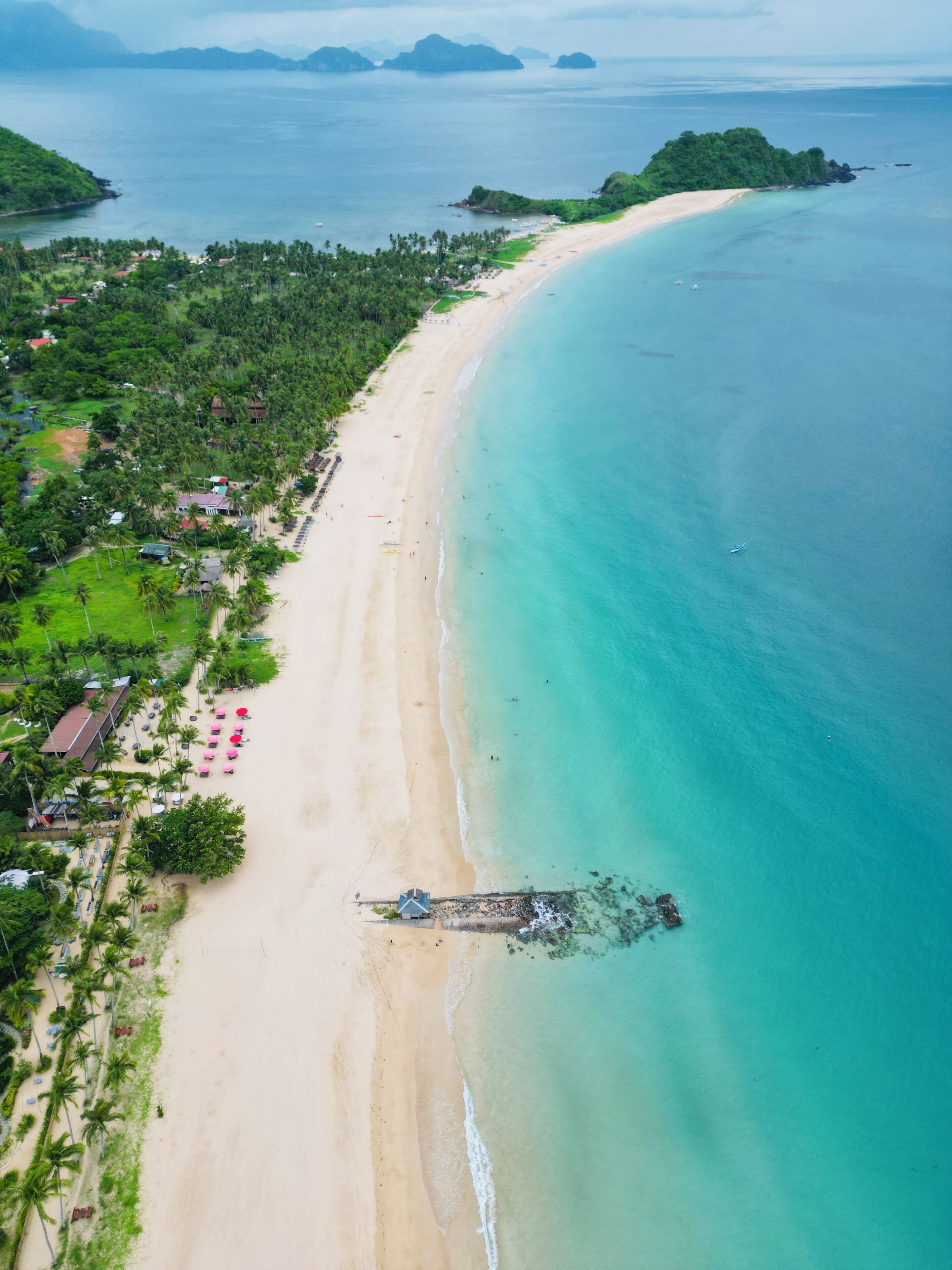 An aerial view of a beach with a clear shoreline, a small pier, and a row of beachside buildings.