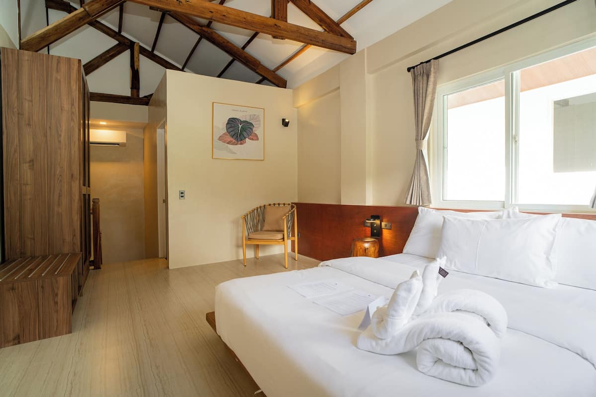Une chambre d’hôtel avec un plafond en bois, un lit avec du linge de lit blanc, une chaise, une lampe fixée au mur et une fenêtre avec des rideaux.