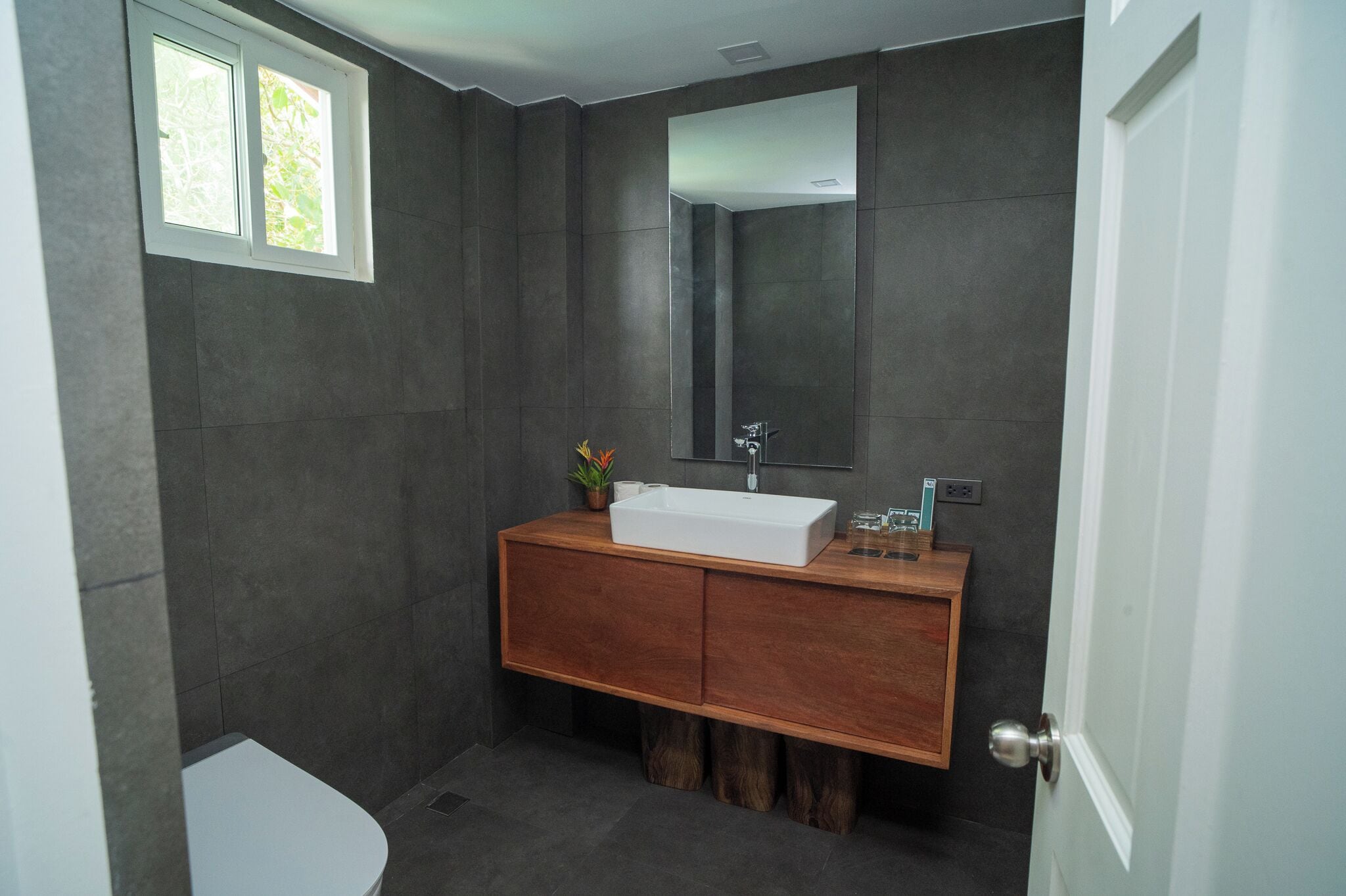 Un baño con lavamanos blanco, paredes de azulejos oscuros, tocador de madera, un espejo y una ventana.