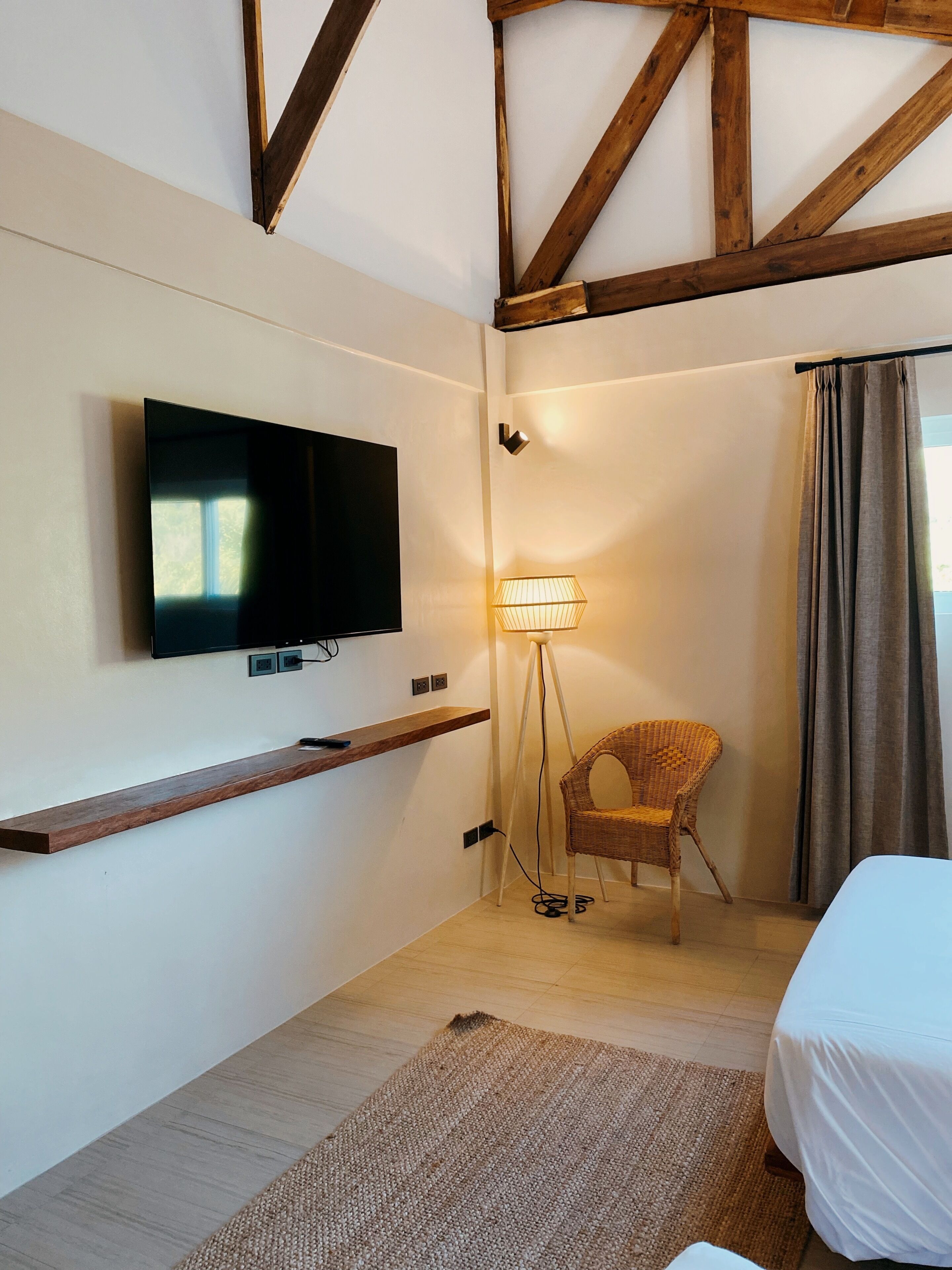 Habitación de hotel con televisión montada, lámpara de pie, silla de mimbre, cama y estructura de vigas de madera en el techo.