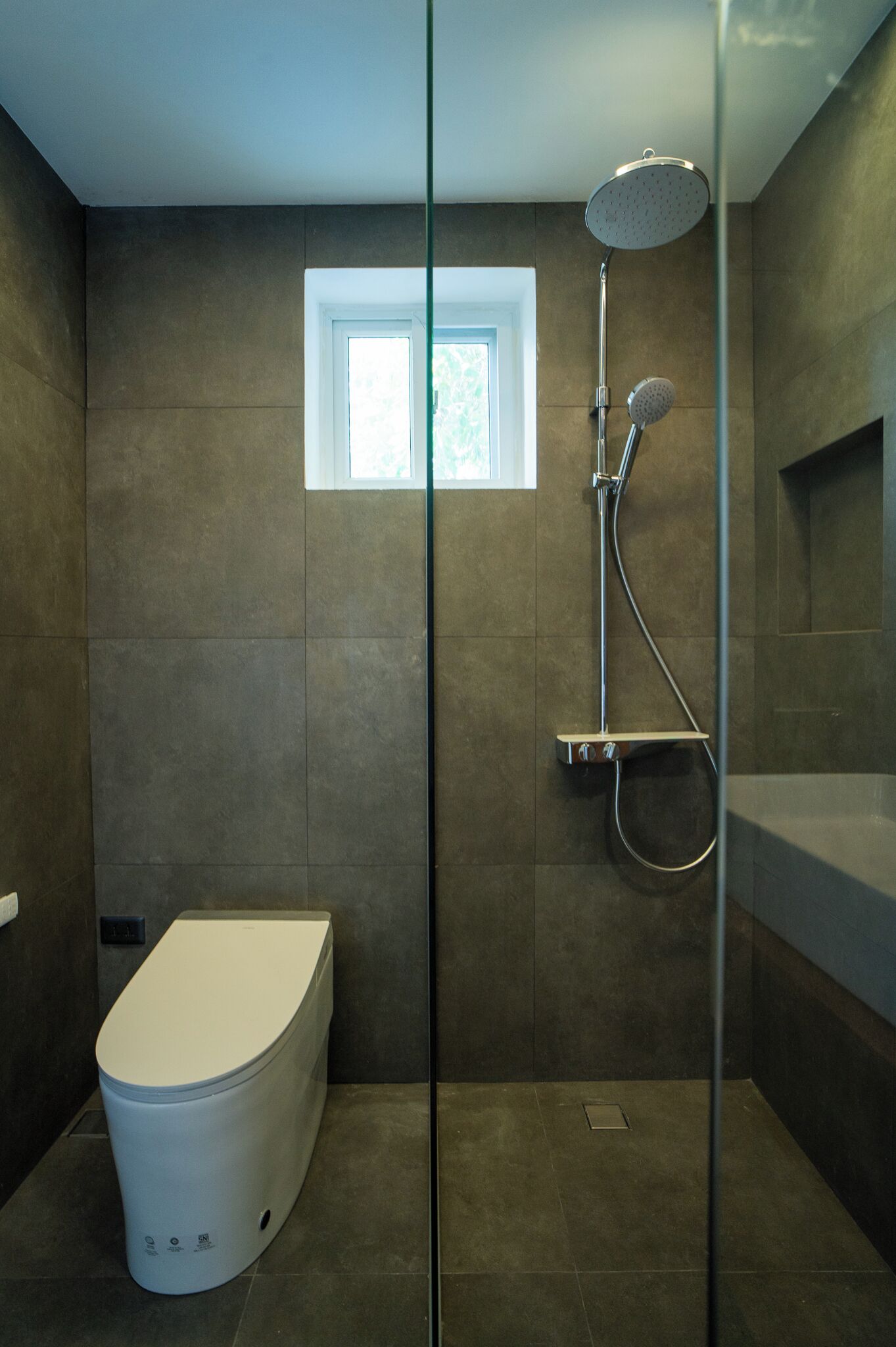 A modern bathroom with a glass-enclosed shower, a toilet, and a window.
