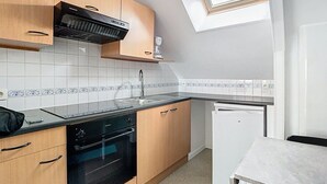 A compact kitchen with wooden cabinets, a black oven, a dishwasher, and a small white refrigerator. - The Little Rest - 40 m² Apartment center of Vitry (Vitry-en-Artois)