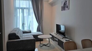 A modern living room with a sofa, a TV, a small round table, and a wicker chair. - BaySuites by Alawa Homes (Kota Kinabalu)