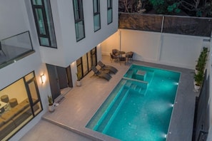 Une piscine extérieure moderne, dotée de chaises longues et d’une table, et entourée d’un bâtiment avec de grandes fenêtres et un balcon.