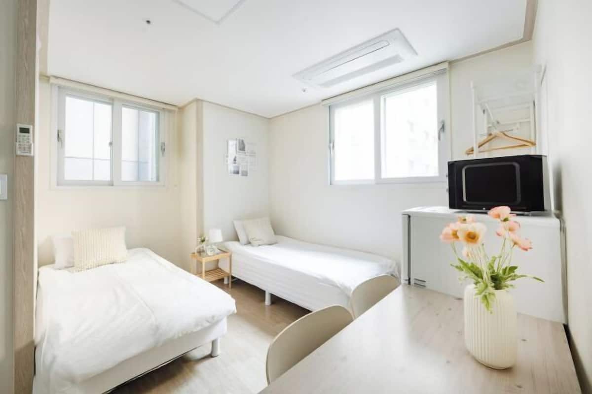 A hotel room with a bed, a desk, a chair, a small table, a TV, and a vase with flowers.