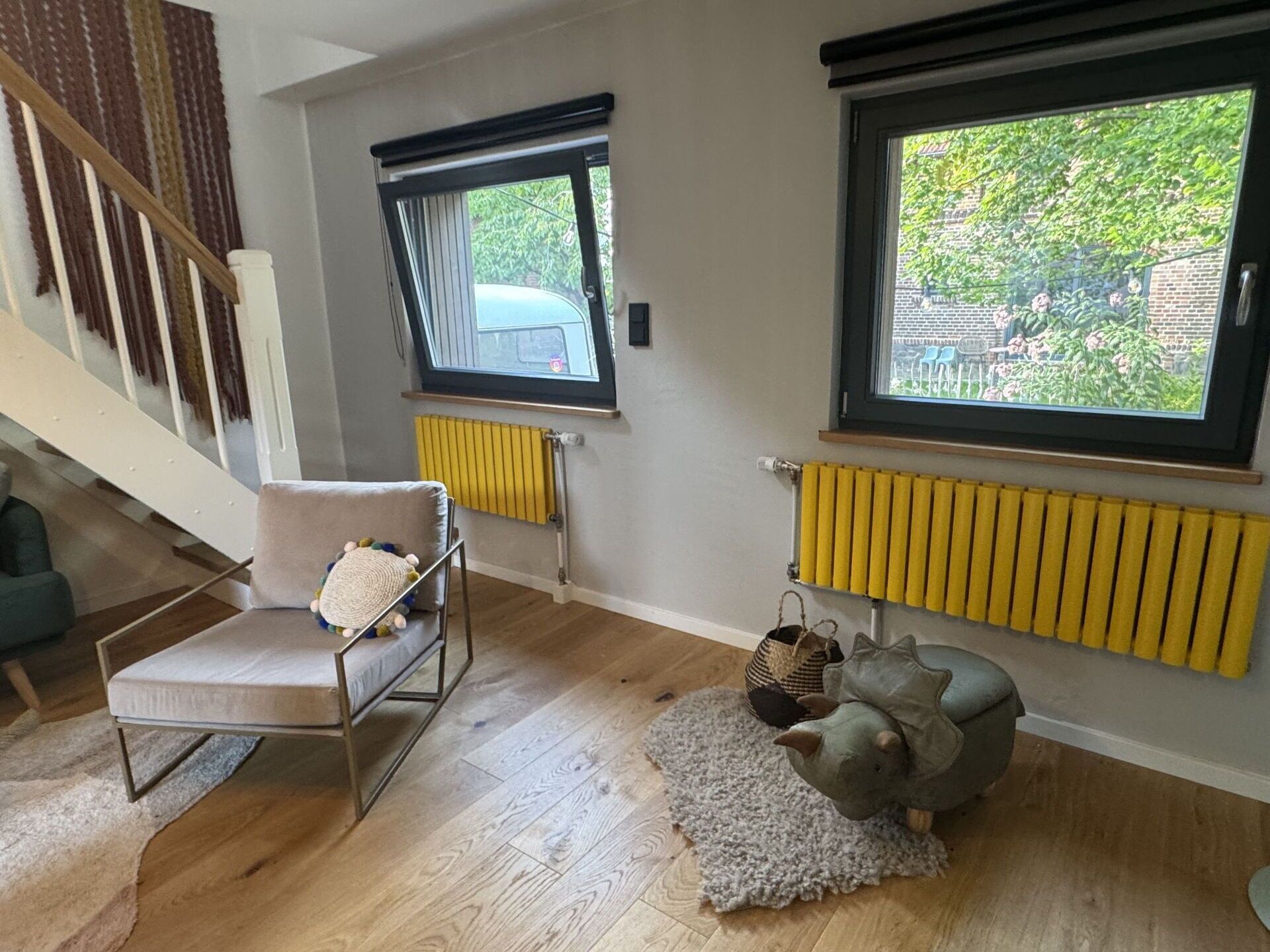 Ein Zimmer mit Treppe, einem Sessel, einer Couch, einem Heizkörper und einem Fenster mit Blick auf Grünflächen.