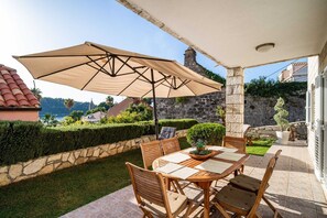 An outdoor patio with a table and chairs, a large umbrella, and a view of the sea and buildings in the background.