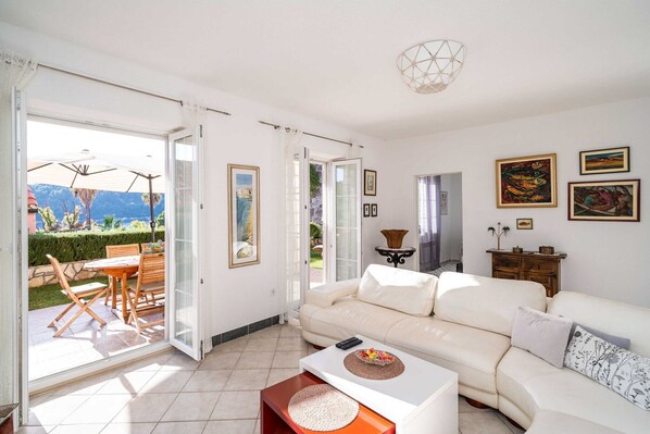 A living room with a white sofa, a coffee table, and a dining area with wooden chairs and a table. There are open doors leading to a balcony with a view of mountains.
