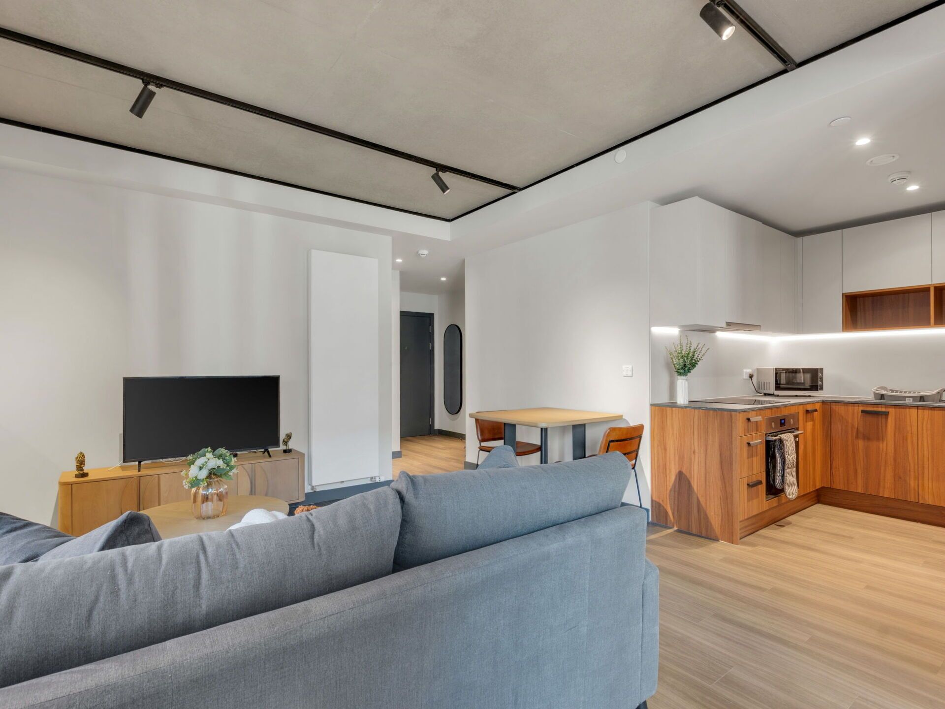 Een moderne woonkamer met een grijze bank, een flatscreen televisie en een eethoek met een houten tafel en stoelen.