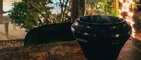A black ceramic cup on a wooden table outdoors with greenery and string lights in the background.