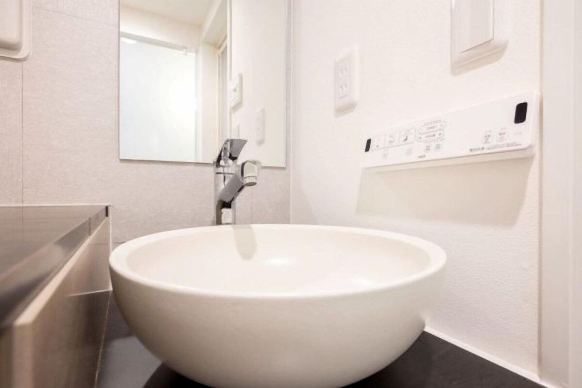 A modern bathroom with a large white sink, a mirror, and a wall-mounted control panel.