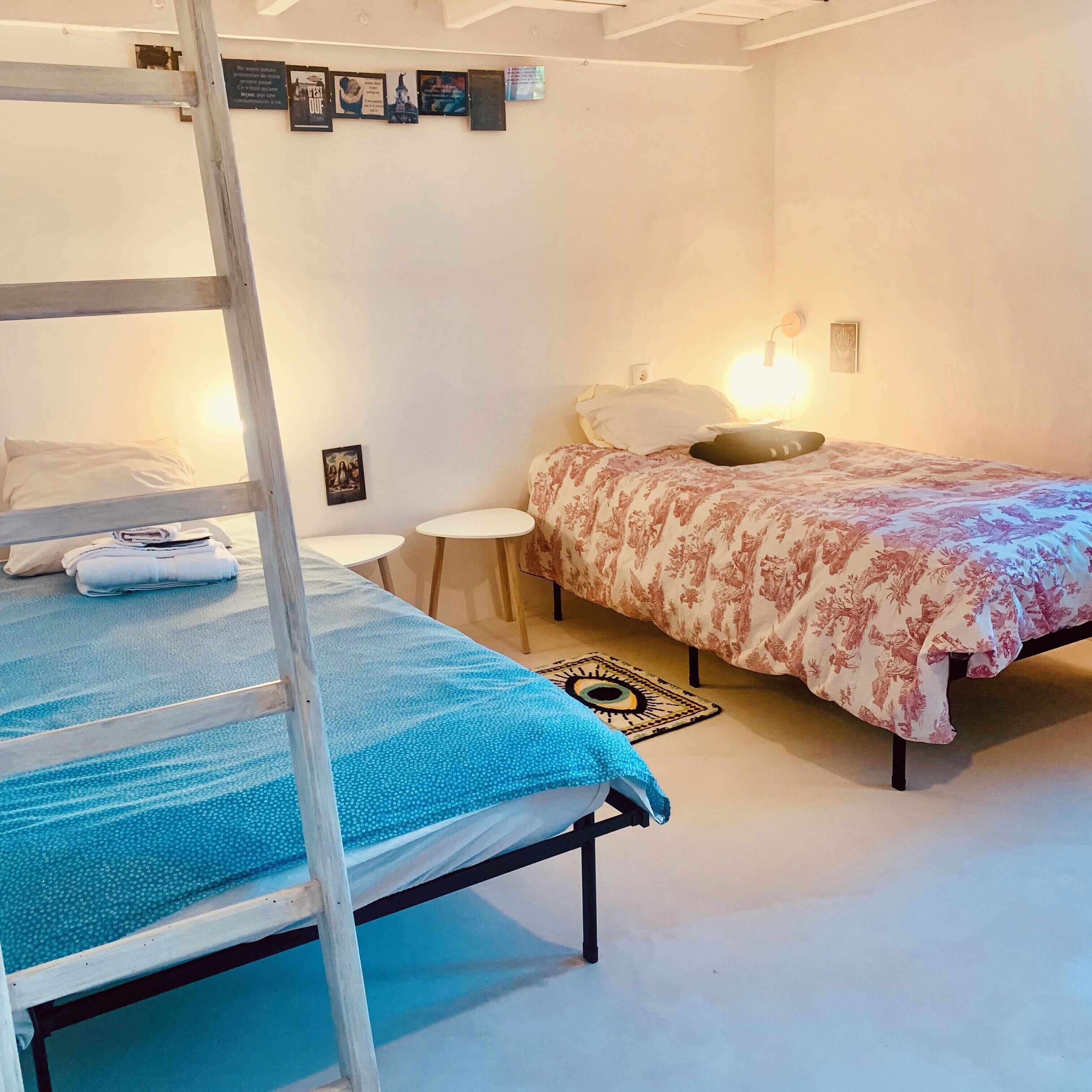 A room with a bunk bed, a single bed, a small table, and a wall-mounted shelf with pictures.