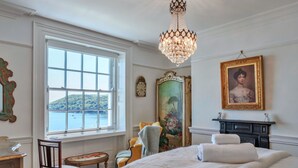 A bedroom with a large window, a chandelier, a bed with white linens, a chair, a small table, and a painting on the wall. - Beautiful  villa for 10 people with WIFI, terrace and pets allowed (Cawsand)