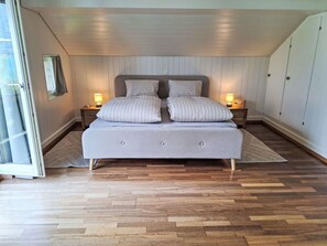 Une chambre à coucher avec un lit, deux tables de chevet avec des lampes, un plancher en bois et une vue sur l’extérieur, visible à travers une porte vitrée.