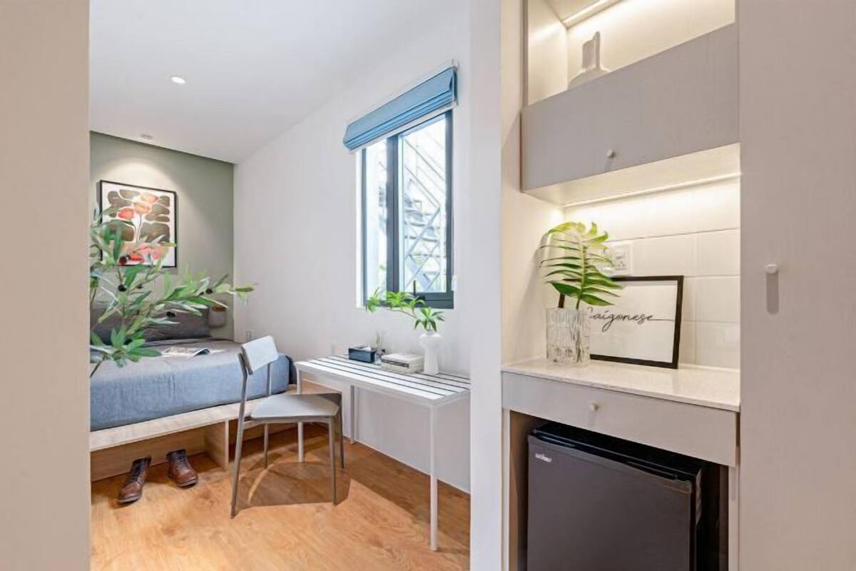 A compact room with a bed, a desk, a chair, a small fridge, and a window with a view of a building.