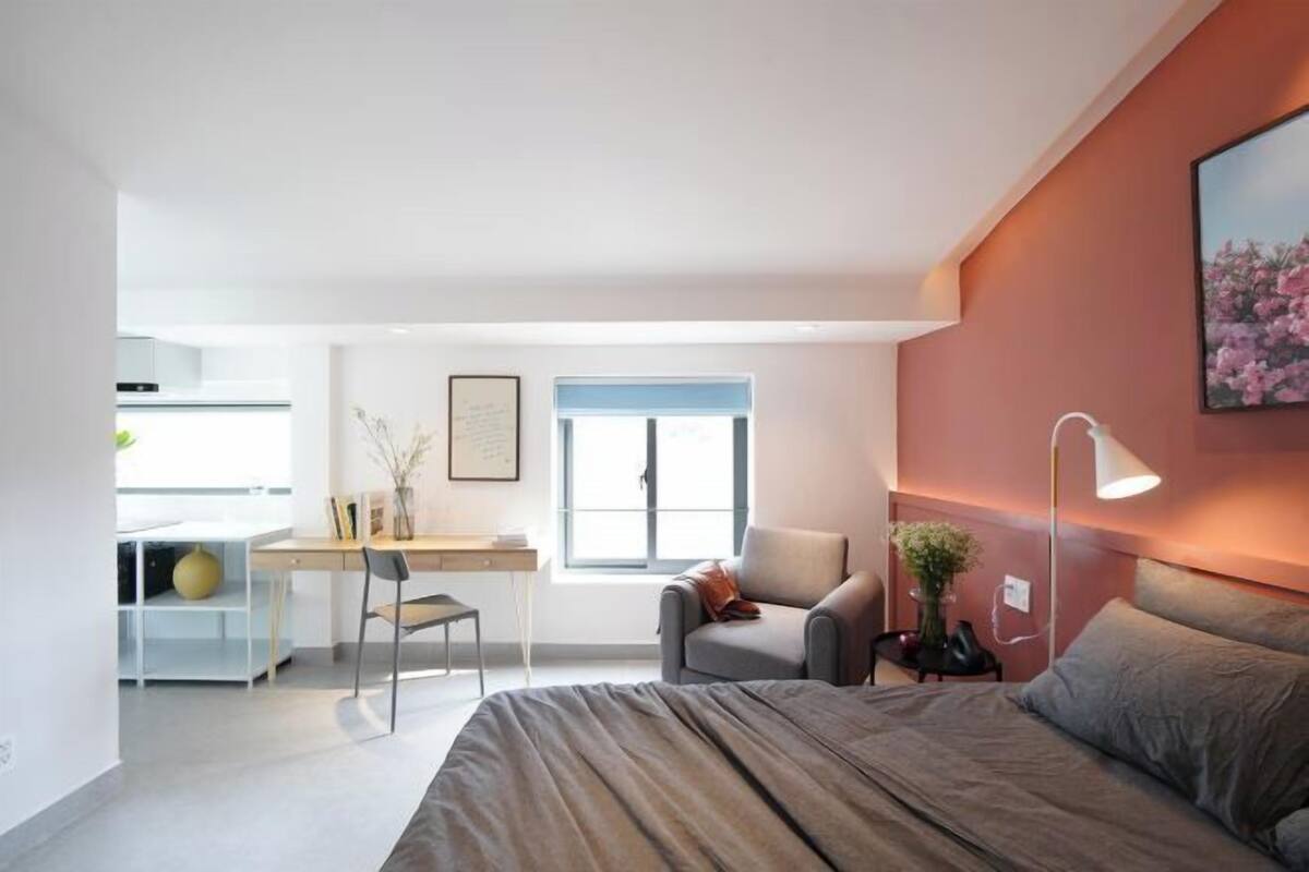 A modern bedroom with a bed, a desk, and a chair, a small table with a lamp, and a window.