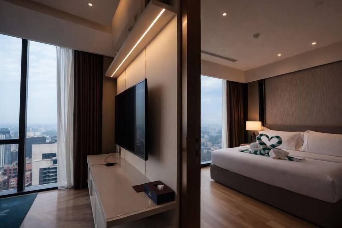 A modern hotel room with a large bed, a flat-screen TV mounted on the wall, a desk with a laptop, and a large window offering a city view.