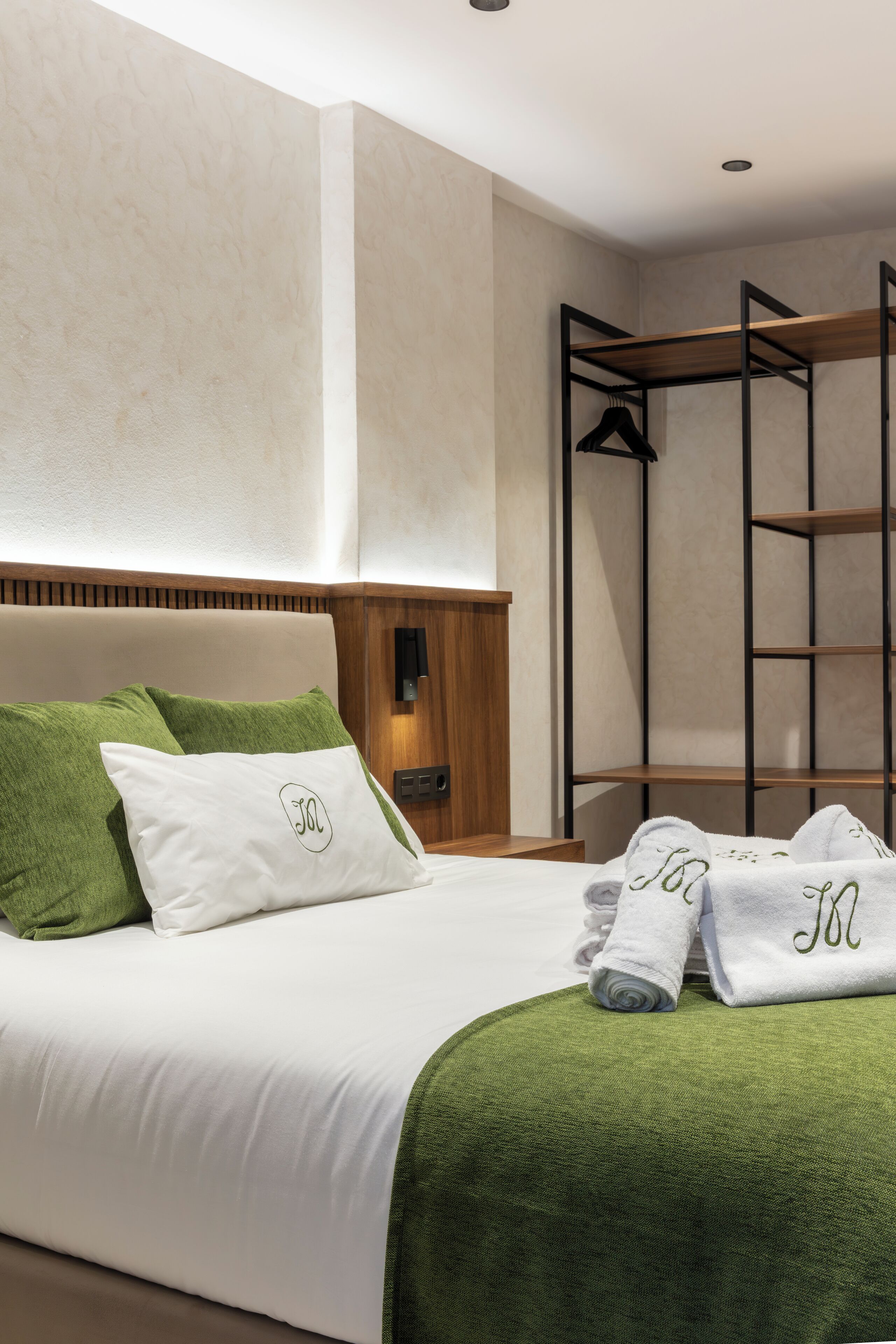 A neatly made bed with white bedding and green pillows, a wooden headboard, and a small wooden shelf unit beside it.