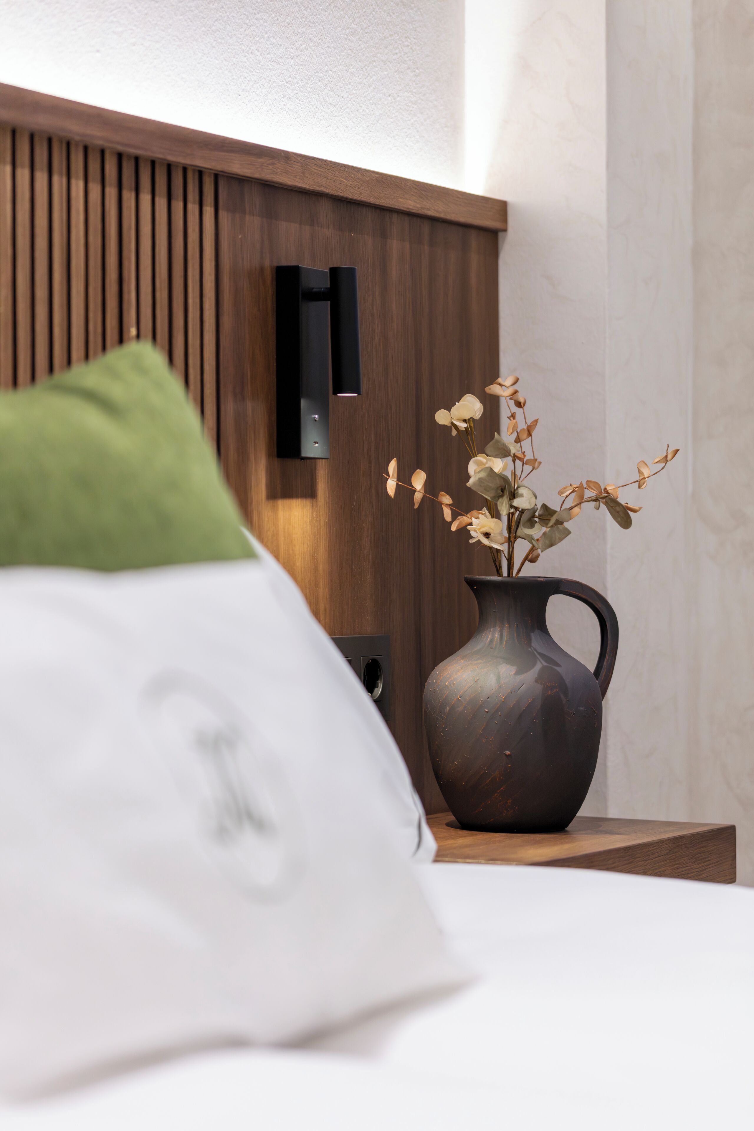 Una cama con un almohadón blanco y uno verde, un jarrón oscuro con flores secas, y un cabecero de madera con un interruptor de luz.