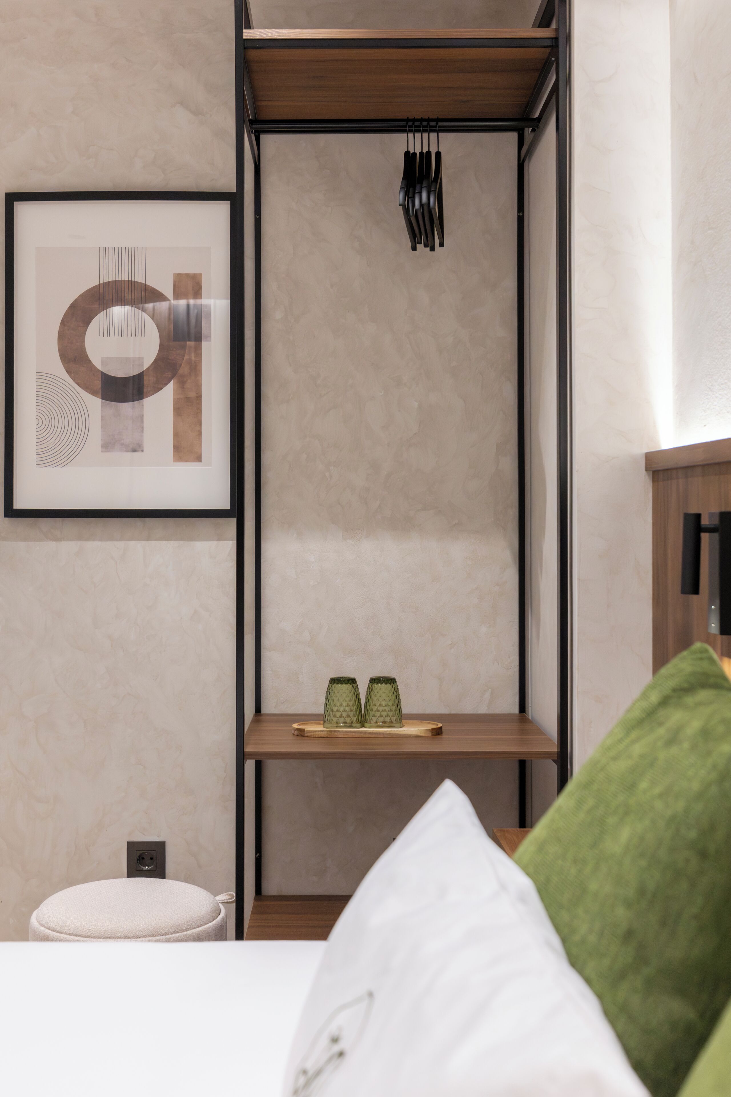 A modern room with a bed, a framed artwork, a shelf with decorative items, and a small stool.