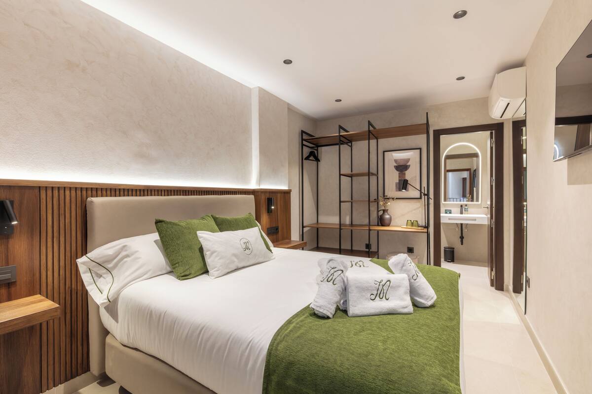 Una habitación de hotel moderna con una cama grande, un plaid verde y un cabecero de madera. Hay un tocador con espejo y lavamanos, además de un pequeño rincón de descanso con una mesa y sillas.