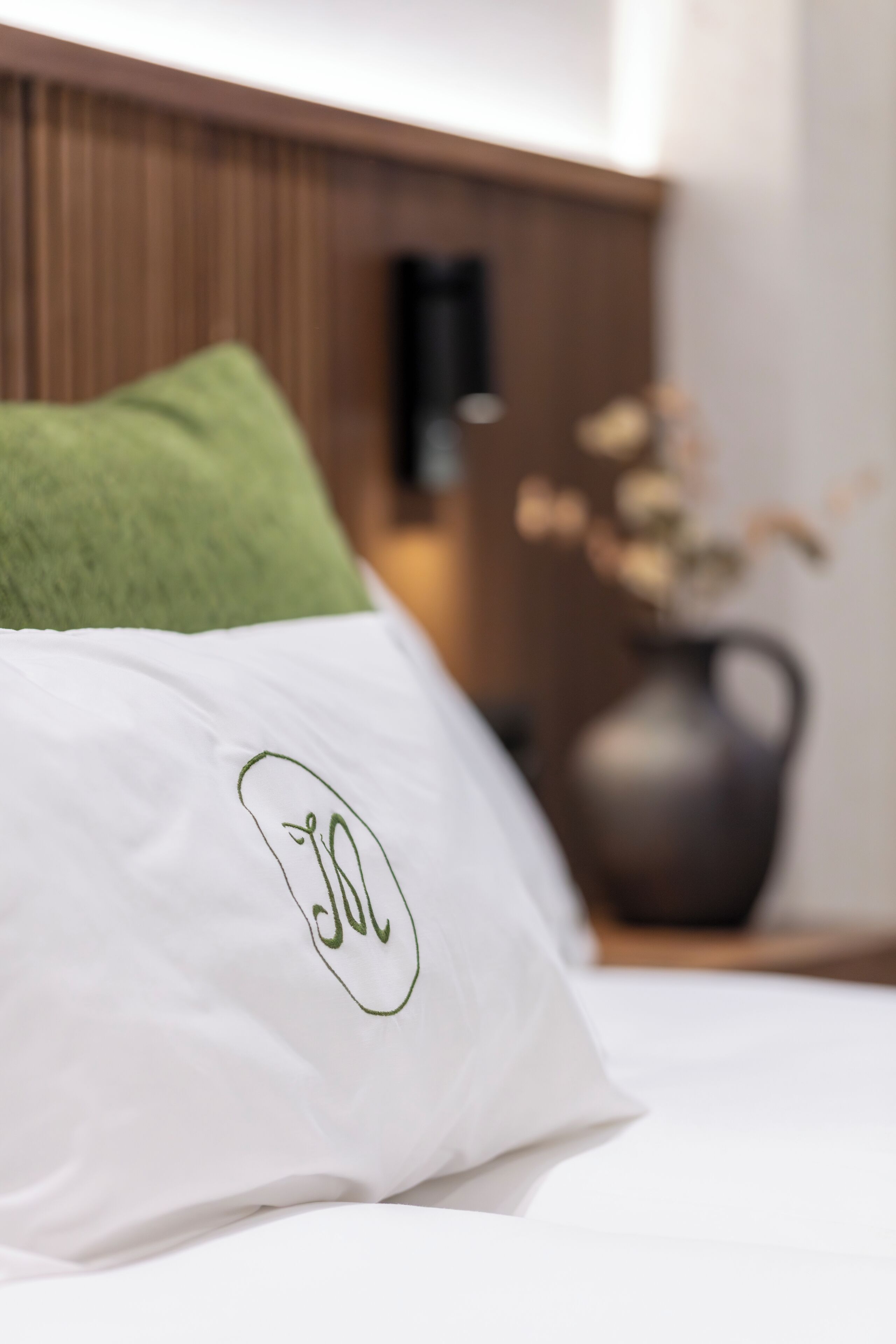A bed with a white pillow featuring a green embroidered logo, a green pillow, and a vase with dried flowers on a wooden nightstand.
