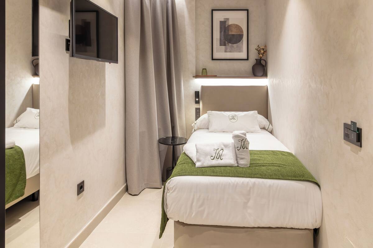 Habitación de hotel con cama, mesita de noche, televisor, estante en la pared y ventana con cortinas.