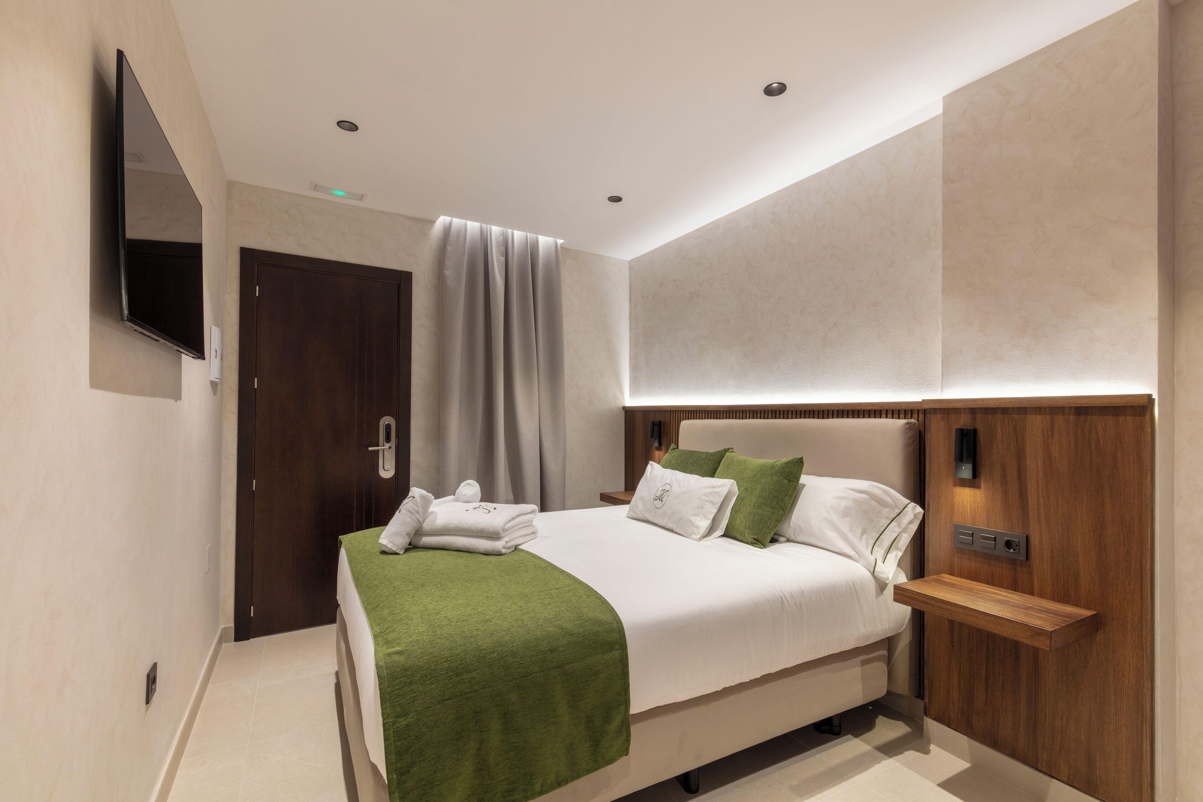A modern hotel room with a large bed, a flat-screen TV mounted on the wall, a wooden nightstand, and a built-in shelf.