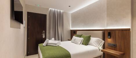 A modern hotel room with a large bed, a flat-screen TV mounted on the wall, a wooden nightstand, and a built-in shelf.