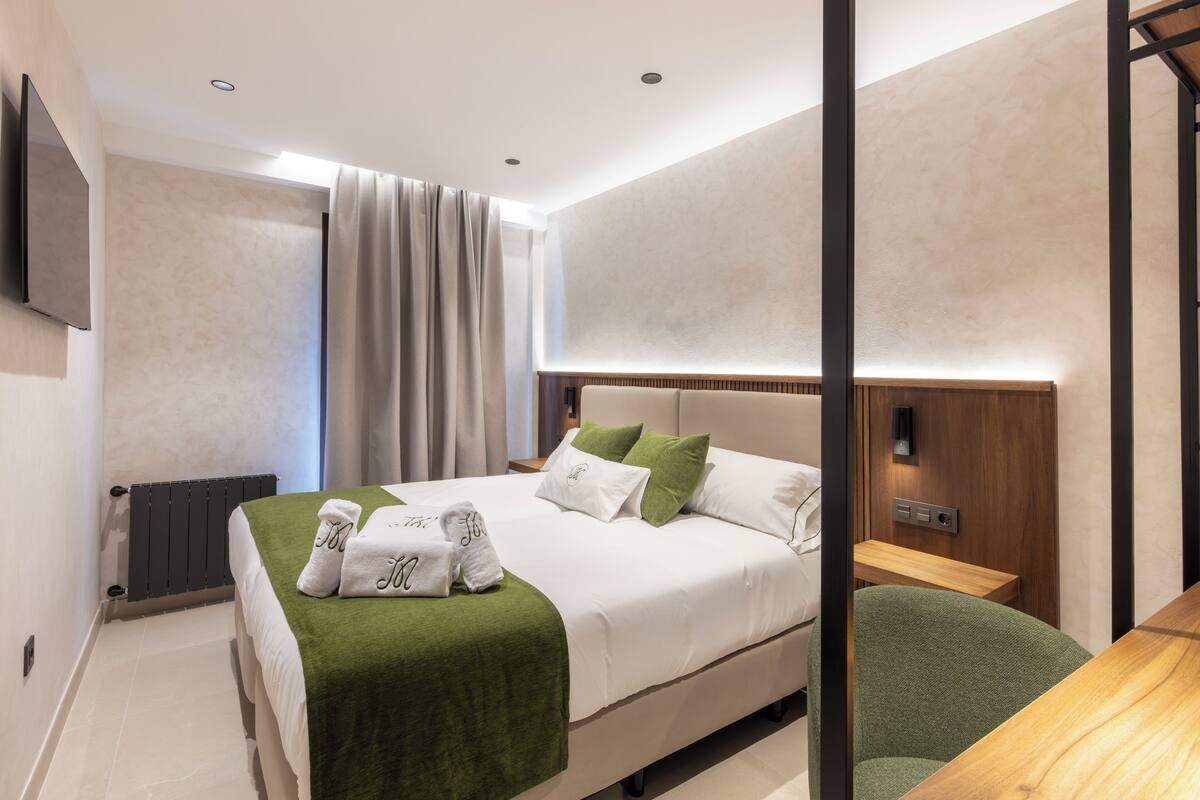 Una habitación de hotel moderna con una cama grande, almohadones verdes, una mesita de noche de madera y un televisor de pantalla plana montado en la pared.