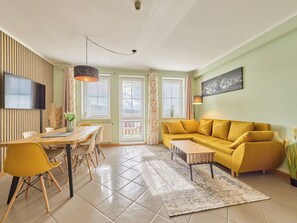 Een moderne woonkamer met een gele bank, een eettafel met stoelen, en een aan de muur bevestigde televisie.