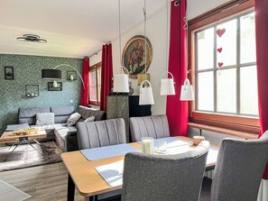 Ein gemütliches Wohnzimmer mit einer Couch, einem Esstisch mit Stühlen, einem Couchtisch und einem großen Fenster mit roten Vorhängen.