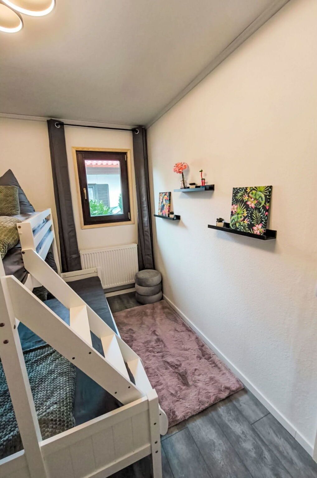 Un pasillo estrecho con una escalera blanca, un radiador, un ventanal con cortinas y dos repisas con objetos decorativos.