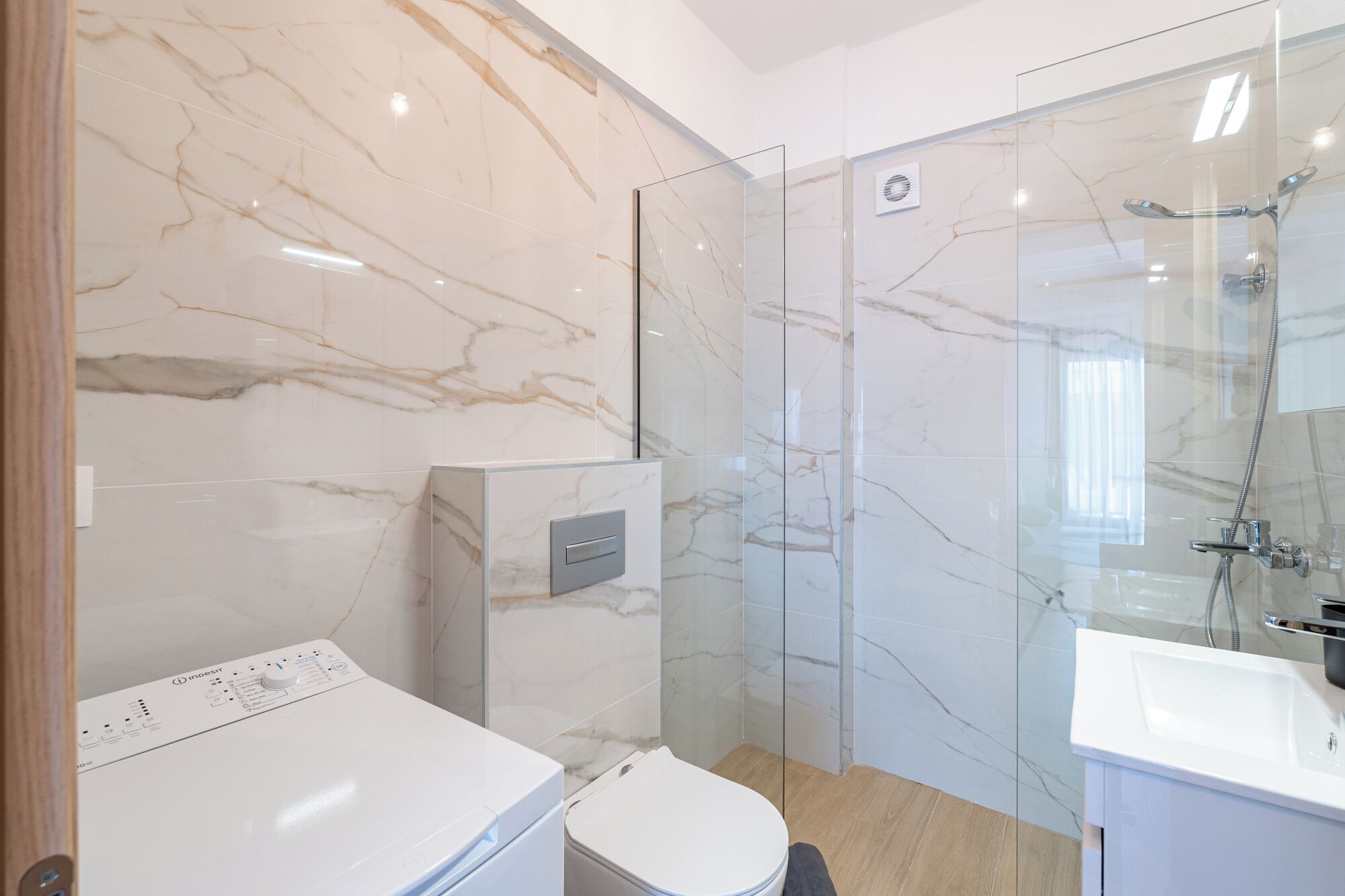Un baño moderno con pared de mármol, cabina de ducha de vidrio, lavadora integrada y lavamanos blanco.