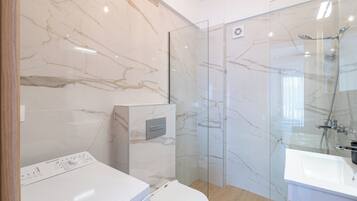 Un baño moderno con pared de mármol, cabina de ducha de vidrio, lavadora integrada y lavamanos blanco.